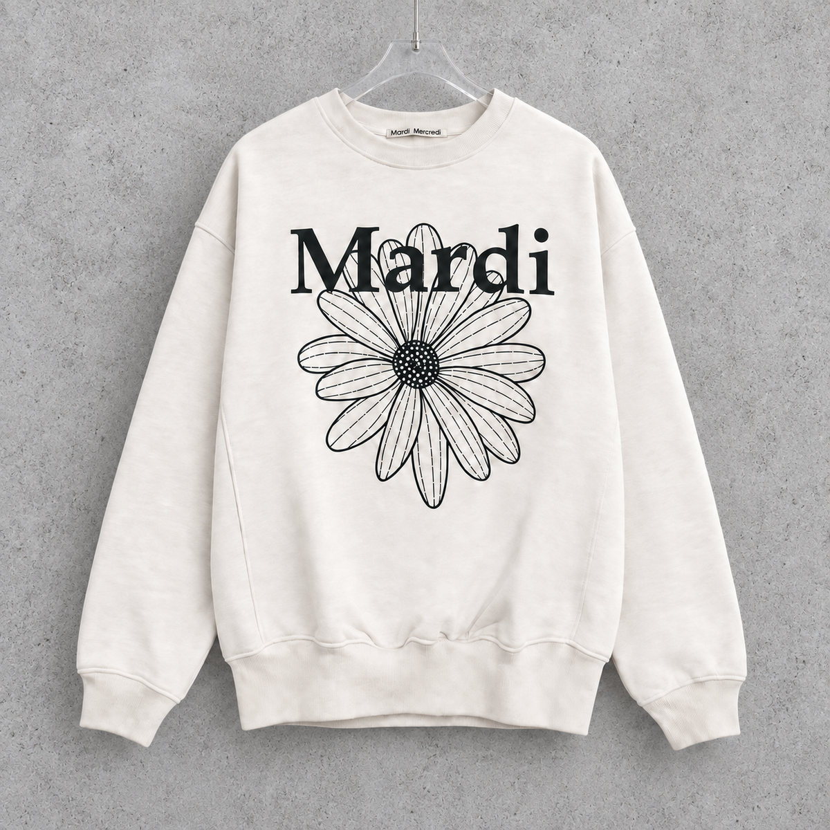 Mardi Mercrediのアイボリーのフラワースウェット。黒いロゴと花柄を正面に見せ、透明ハンガーとコンクリート背景で撮影風に合成した画像