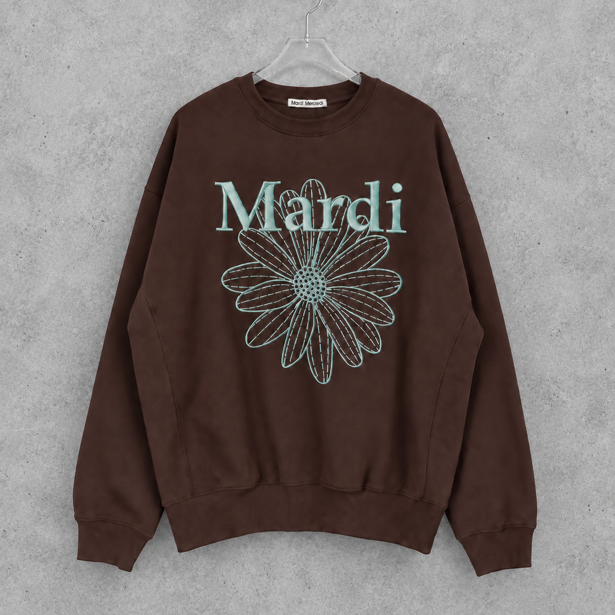 Mardi Mercrediのブラウンのフラワースウェット。淡いブルーのロゴと花柄を正面に見せ、透明ハンガーとコンクリート背景で撮影風に合成した画像